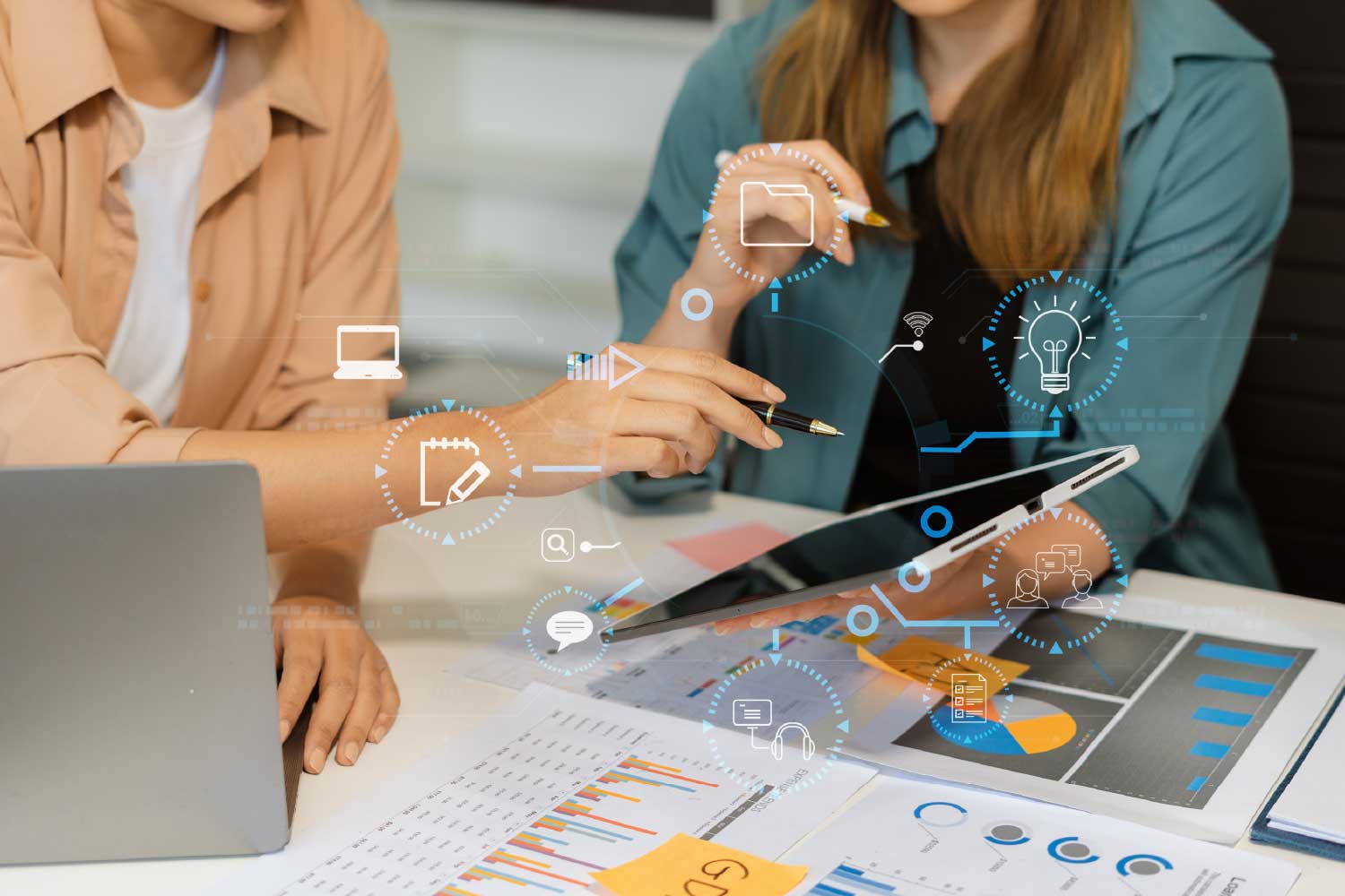 Two individuals collaborate with a tablet at a desk filled with documents