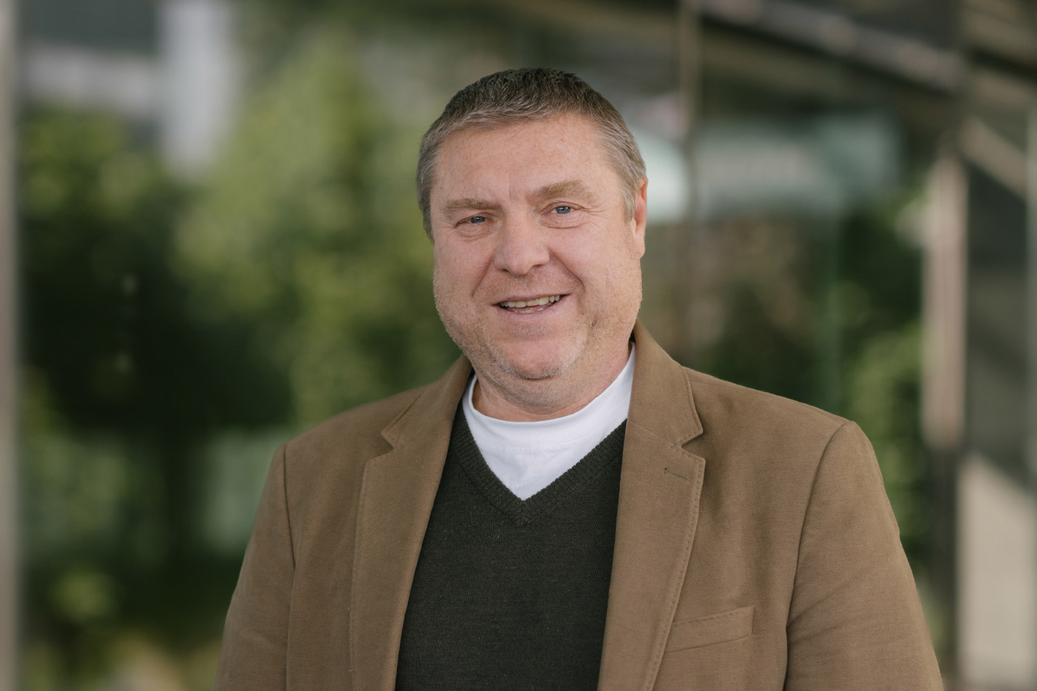Headshot of Matt Neil wearing a black v-neck jumper and brown blazer standing outside.