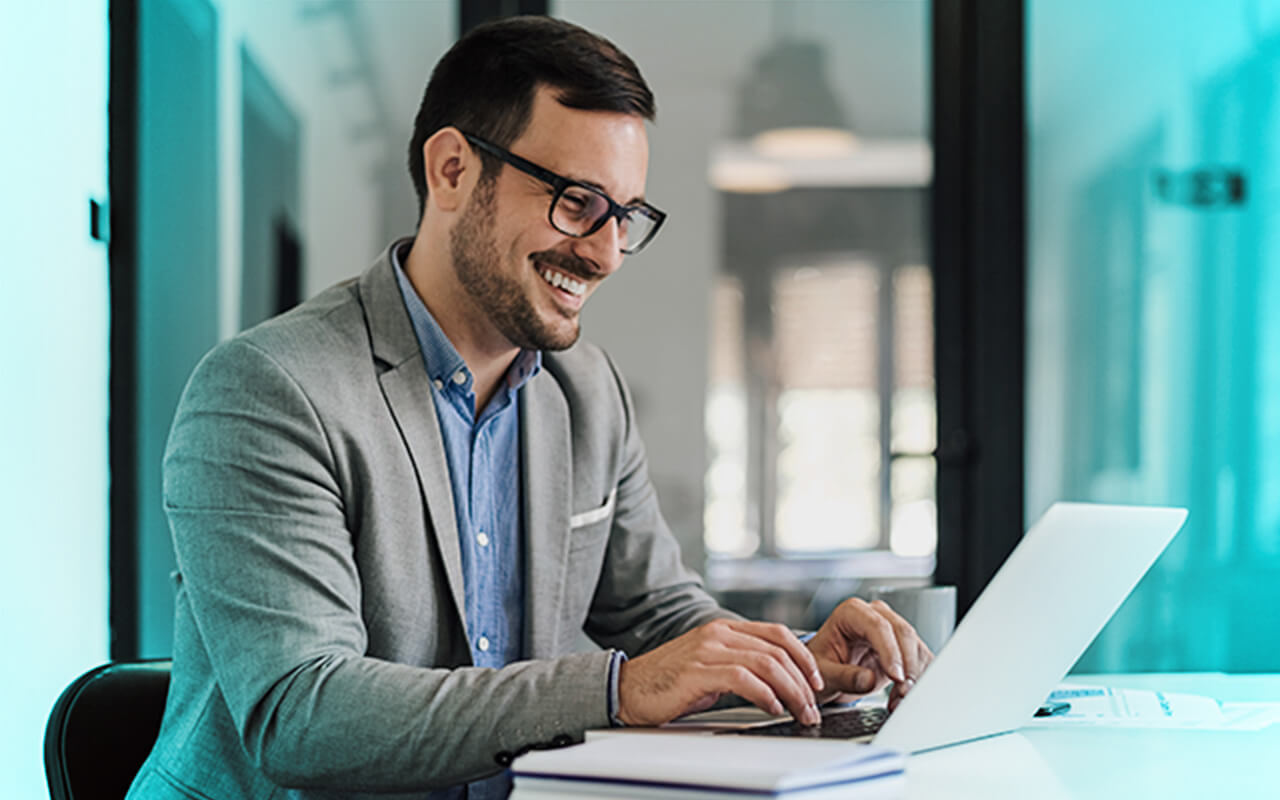 A smiling young professional working on a laptop at a desk in a modern office.
