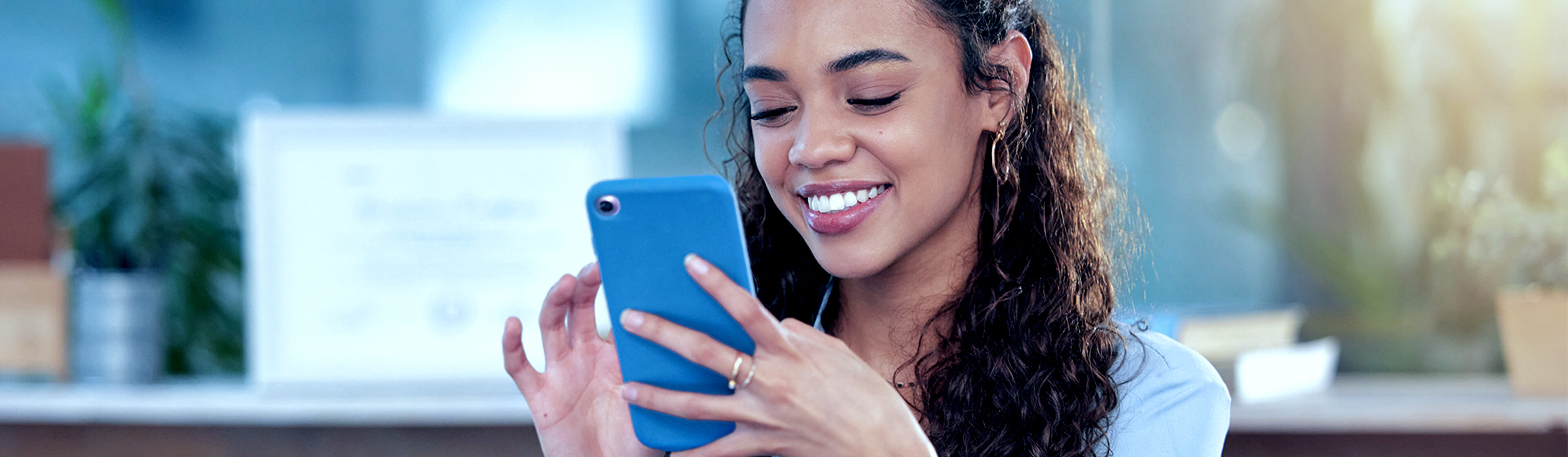 A woman wearing a sky blue coloured shirt smiling while looking at a mobile phone