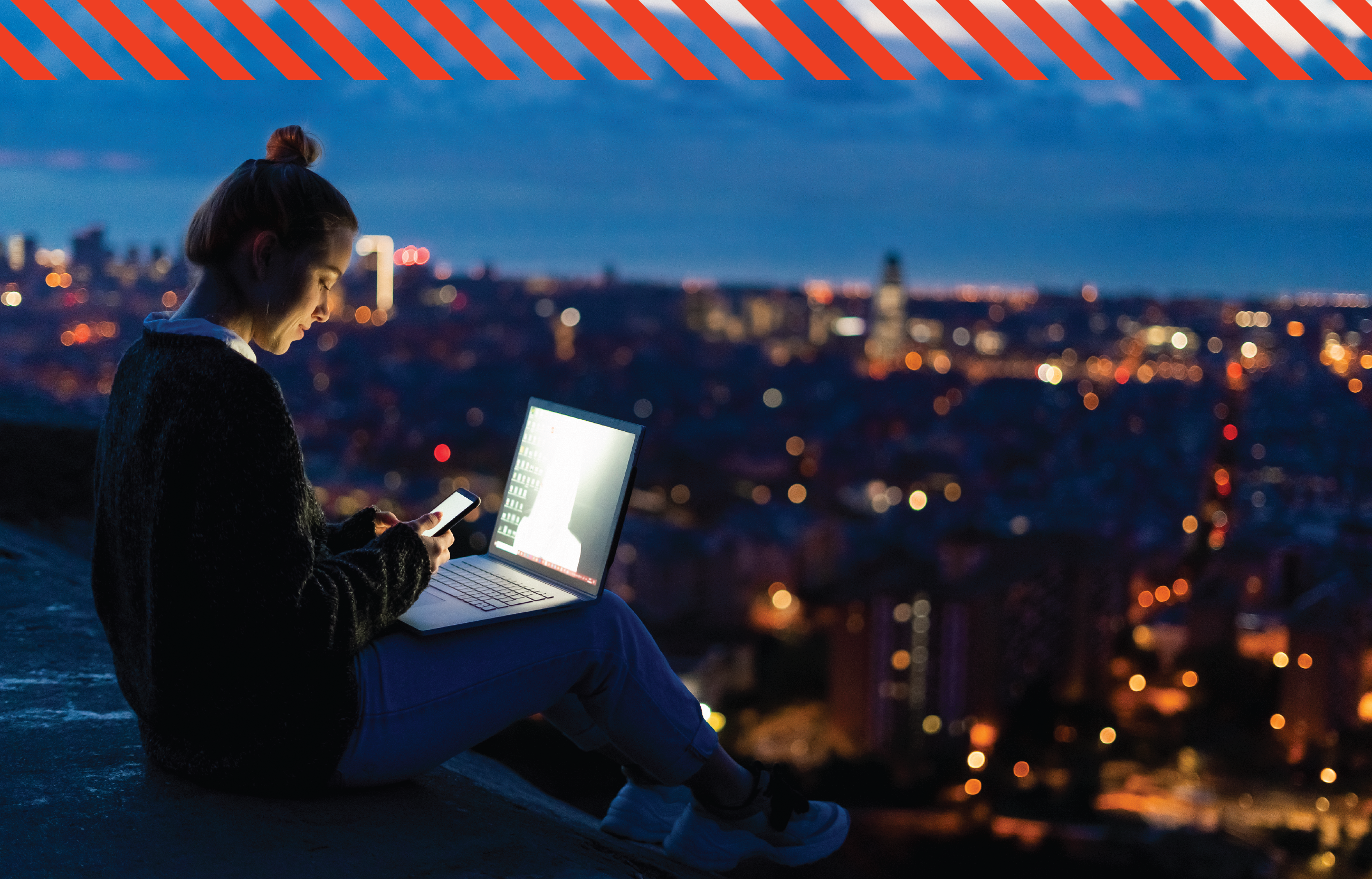 A woman looking over a city in the dark whilst using her phone and laptop
